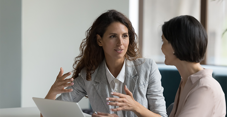 two-business-women-sit-desk-discuss